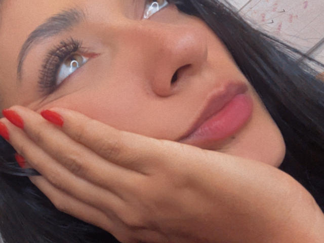 AmeliRoux's face, with dark hair, red nails, and hand on cheek, gazing up with blue eyes, pink lips, and slightly upturned nose, in a soft, natural light.