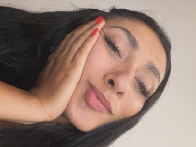 AmeliRoux a la peau mate, de longs cheveux noirs, des ongles rouges, et elle sourit, sa main sur son visage, mettant en valeur ses cils et ses lèvres roses..