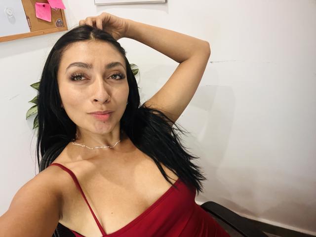 AmeliRoux with long black hair, red dress, and a hand in her hair, showing off her tits and a slight pout. She's wearing a silver necklace.