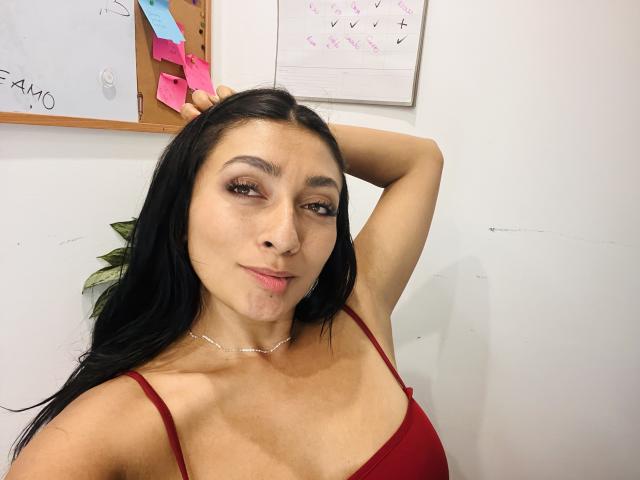 AmeliRoux with long black hair, wearing a red spaghetti-strap top, hand behind head, slight smile, visible boob cleavage, brown eyes, light skin, small nose ring, white wall background.