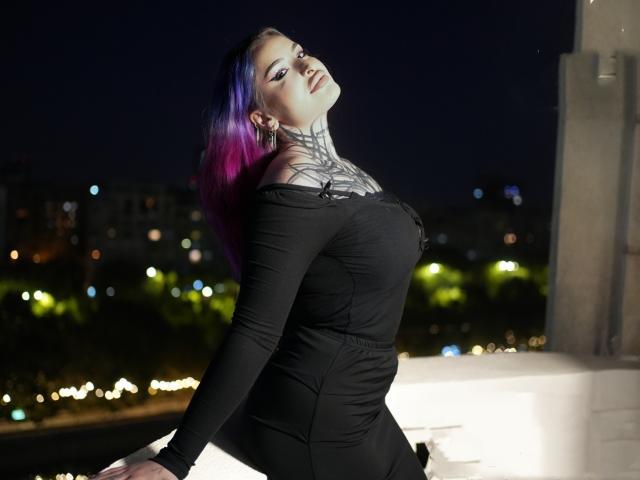 ElviraRae mit lila und blauen Haaren, einem schwarzen, schulterfreien Kleid, einem tätowierten Hals, die sich zurücklehnt und ihre Brüste zeigt, in einer nächtlichen Stadtkulisse, mit leicht geöffnetem Mund..