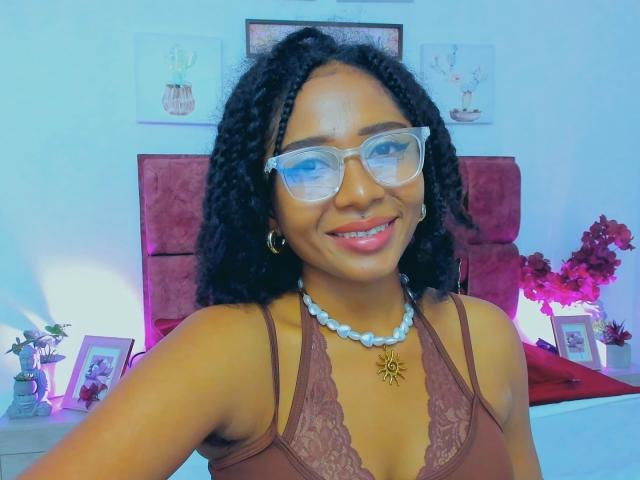 Megan Williams, avec ses cheveux noirs bouclés, ses lunettes, son soutien-gorge en dentelle marron, souriante, portant un collier de perles bleues, un pendentif en or, du rouge à lèvres rose et des boucles d'oreilles créoles, assise dans une pièce très éclairée..