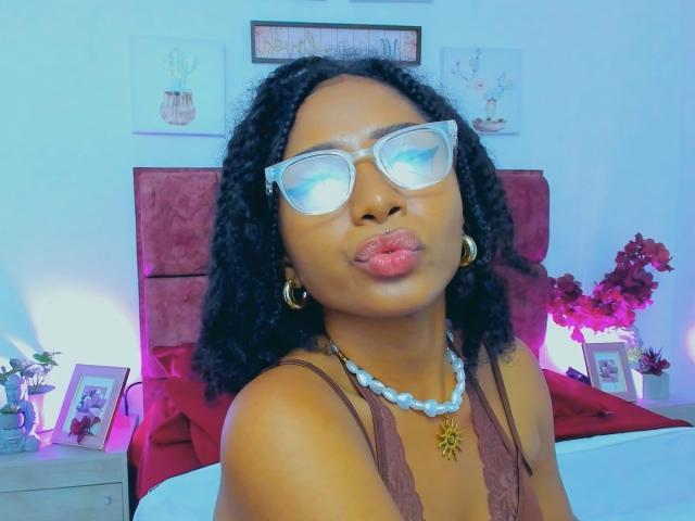 Megan Williams, avec une peau brun foncé, des cheveux noirs bouclés, des lunettes claires, des boucles d'oreilles dorées, un collier de perles blanches, un haut en dentelle marron, des lèvres roses pulpeuses, et un lit rouge derrière elle..