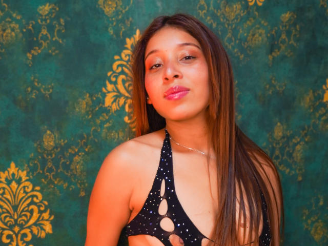 JaneKetty, con cabello largo y castaño, vestida con un top negro de tirantes con diseños recortados, mirando a la cámara con labios rosados ligeramente fruncidos. Su piel es morena y es atractiva..