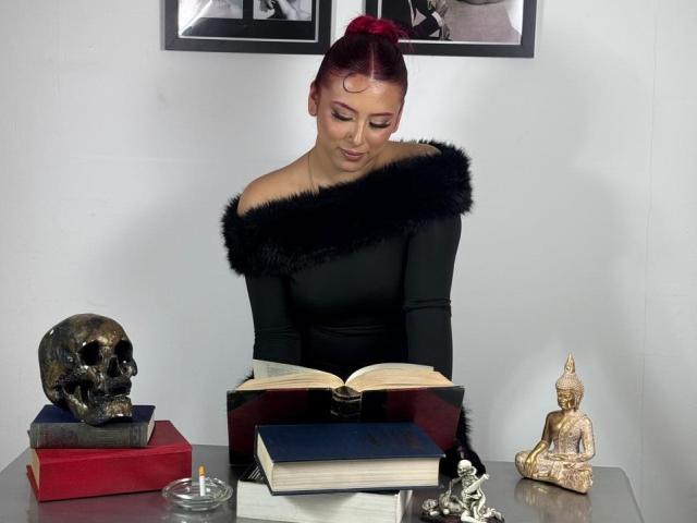 MaddyMills with red hair, black off-shoulder top with fur, reading book, skull on red box, Buddha statue, candle, and books on gray table, looking down.