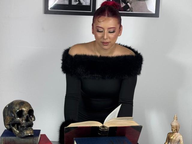 MaddyMills with red hair, black off-shoulder dress with fur trim, reading a book, skull on left, Buddha statue on right, focused, book open in front.