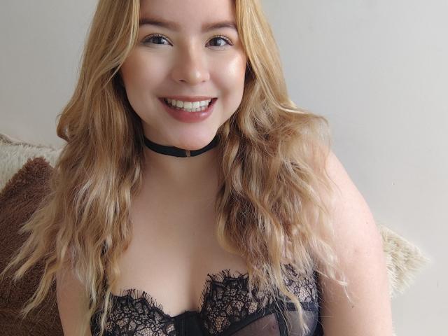 AshleySofia with blonde wavy hair, smiling, wearing a black lace bra and choker, showing her tits, with light skin and cute face, in a selfie.