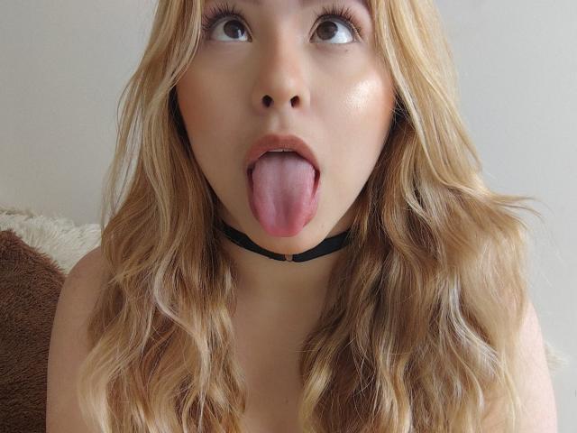 AshleySofia, con i capelli biondi e ondulati, un collare nero e la lingua fuori, guarda in alto con grandi occhi marroni, il naso leggermente rivolto verso l'alto e la bocca aperta..