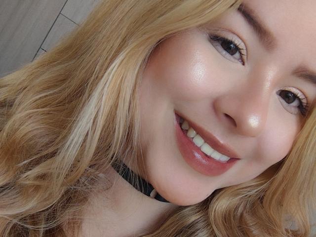 AshleySofia's smiling face with glossy pink lips, shiny skin, and blonde wavy hair, wearing a black choker, has cute white teeth and brown eyes.