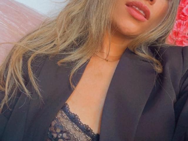 AdharaCooperr, com longos cabelos loiros ondulados, vestindo um blazer preto, com um sutiã de renda preta à mostra, exibindo seus seios. Ela tem lábios rosados e um colar de ouro..