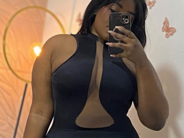 Nicol aus Zürich, mit dunkler Haut, schwarzen Haaren, trägt ein schwarzes, figurbetontes, ärmelloses Kleid, hält ein Telefon, bedeckt ihr Gesicht, wobei ihre großen, runden Brüste und eine sichtbare Bräune zu sehen sind..