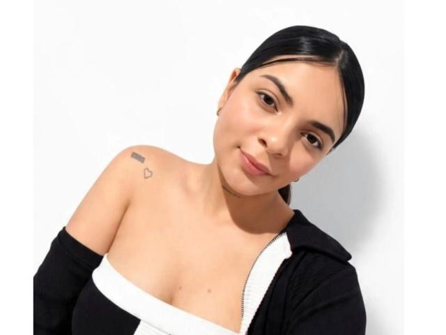 ArantaRossi has medium tits, a small heart tattoo on the left shoulder, and is wearing a black and white off-shoulder top, with black hair and a slight smile.