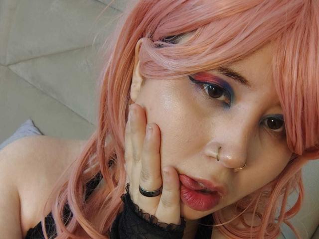 WendyMonet with pink hair, blue and pink eyeshadow, nose ring, and black lace finger, touching her face, looking up, with parted lips, showing her pink nipples.