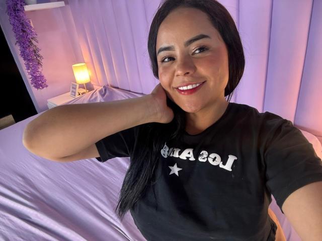 NixieRose with dark hair, smiling, wearing a black shirt, touching her neck, purple bed, white star on shirt, lamp, purple curtain, and purple garland in the background.