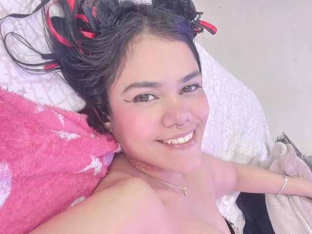 TarahDustin con capelli neri raccolti in trecce rosse e nere, con un anello al naso, sorridente, su un cuscino rosa soffice, su un letto bianco con motivi, e con un braccialetto d'argento, mostrando le spalle scoperte..