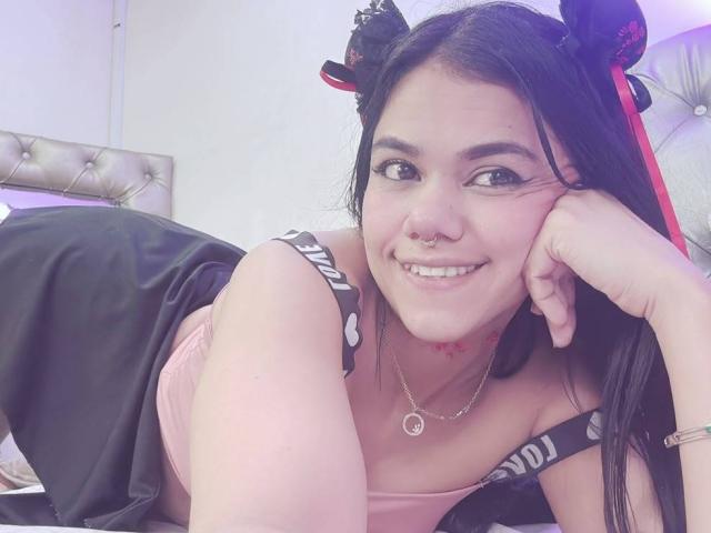 TarahDustin, con i capelli neri, indossa una maglietta rosa e una gonna nera, è sdraiata sul letto, sorride, ha un piercing al naso, un tatuaggio a forma di cuore rosso, una collana rosa, la testa appoggiata sulla mano, è carina, giocosa, sexy..