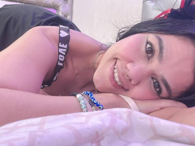 TarahDustin, avec ses cheveux foncés, son anneau de nez et son sourire, est allongée sur un lit, vêtue d'un haut noir avec un bandeau "LOVE", de bracelets et d'un bandeau rose, montrant sa poitrine et son joli visage..