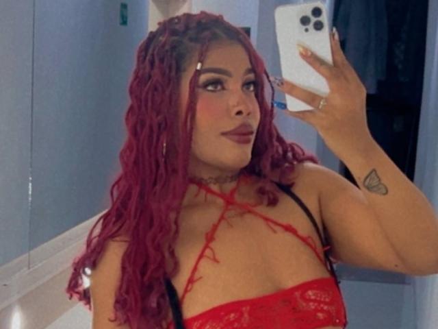 LexieBloom, con cabello rojo trenzado, vistiendo una blusa roja de encaje, tomando una selfie en el espejo, mostrando un tatuaje en su brazo derecho, sosteniendo un iPhone. Sus pechos son pequeños..
