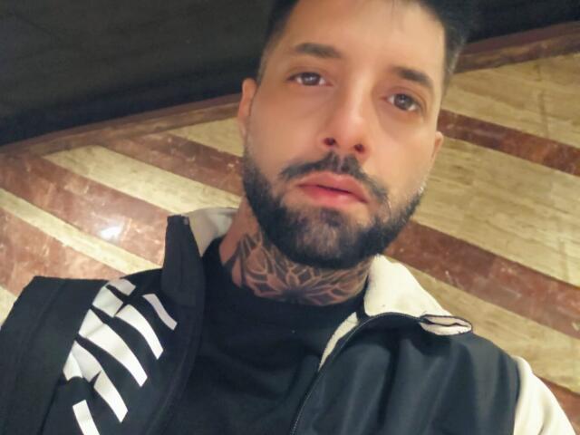 TommyDum a un visage barbu, des tatouages sur le cou et des cheveux noirs courts. Il porte une veste Adidas noire et blanche. Ses yeux marrons semblent légèrement décentrés par rapport à la caméra..