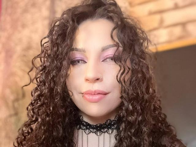ScarlattaaDesire, avec ses cheveux bruns bouclés, son maquillage rose sur les paupières, un anneau au nez, un collier en dentelle noire et du rouge à lèvres rose, regarde directement l'appareil photo avec un léger sourire..