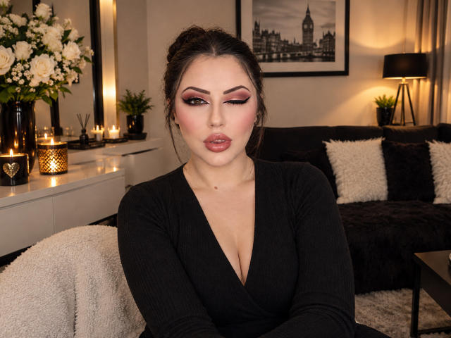 MssBlackWidow with dark hair in a bun, wearing a black V-neck, showing off her tits in a cozy, lit-up room with candles and flowers. She's winking seductively.