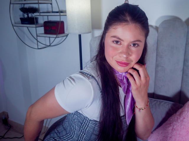 SiennaBeumont con lunghi capelli castani, camicia bianca, vestito a quadretti blu, sciarpa viola, sorridente, mano sul viso, rossetto rosa, luce soffusa, parete bianca, testiera grigia, cuscino rosa, braccialetto..