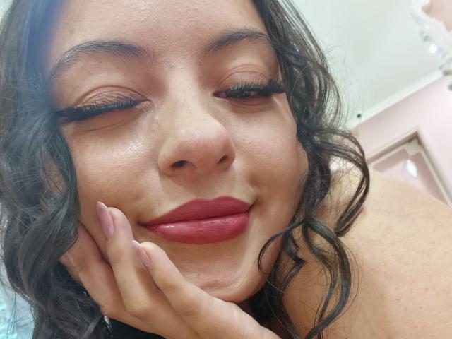 MiiaBennet with winking eyes, red lipstick, and curled black hair, smiling slightly, hand under chin, long eyelashes, pink nails, and soft skin, in a slightly blurry selfie.