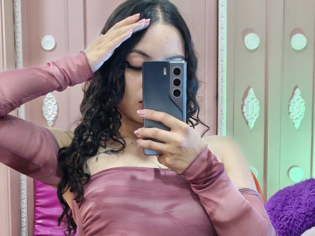 Miia Bennet, con i capelli neri ricci, indossa una maglietta rosa a spalle scoperte, tiene un telefono davanti al viso, con una mano che le copre la fronte, ha le unghie rosa, sullo sfondo c'è un cuscino viola..