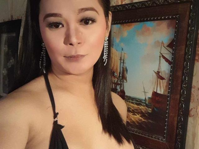 BeautifulWildcock with long black hair, wearing a black halter top, showing her bare shoulders, and large diamond earrings, looking at the camera with a slight smile.