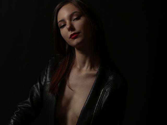 WendyDemmy with dark brown hair, red lipstick, and black leather jacket, exposing small bare breasts, against a black background. She has a confident, slightly smug expression.