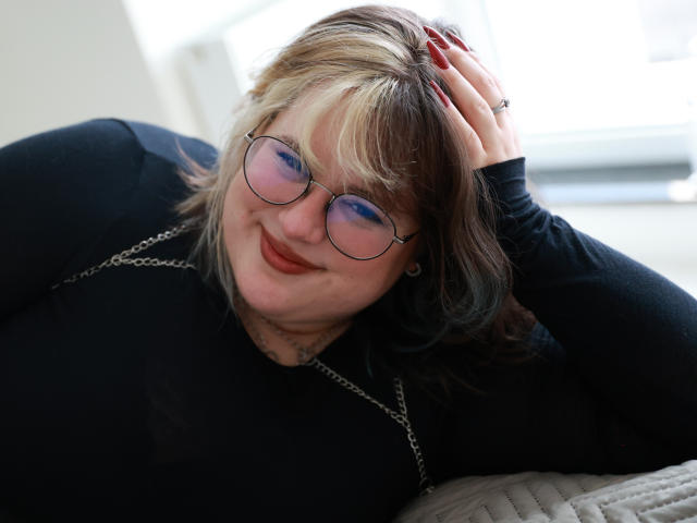 OpheliaLush with blonde and brown hair, glasses, red nails, black top, smiling, hand in hair, wearing a chain necklace, laying on a white surface, looking cute.