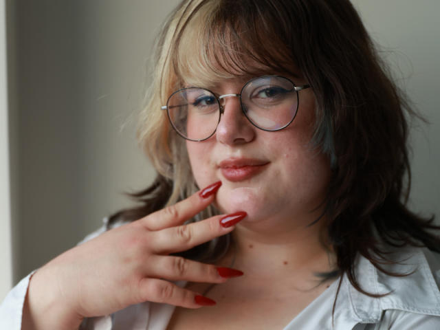 OpheliaLush has pale skin, blonde and brown wavy hair, glasses, red nails, and a subtle smile. She's wearing a white shirt, touching her cheek with her hand.