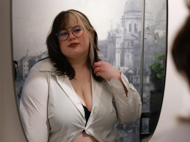 OpheliaLush with glasses, white shirt tied below showing black bra, blonde and black hair, slight smile, hand near neck, reflection in mirror, plus-size, pale skin.