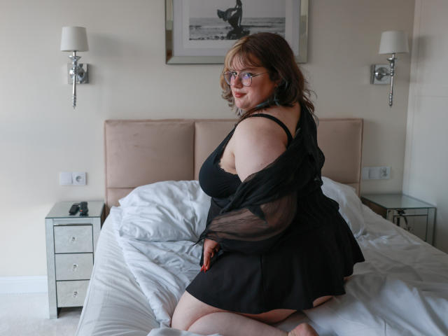 OpheliaLush with glasses, black sheer dress, exposing her tits and ass, kneeling on a bed, looking over her shoulder, smiling, in a bedroom with wall sconces.