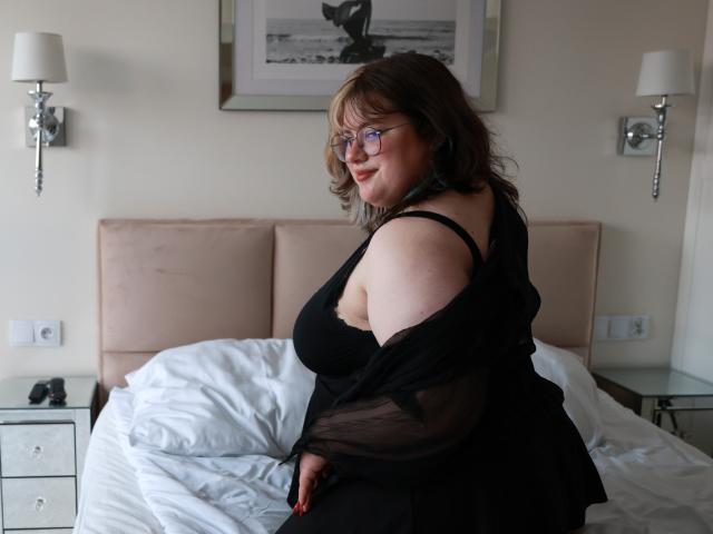 OpheliaLush, a curvy woman with glasses and brown hair, sits on a bed, wearing a black sheer top, exposing her large, round tits and smiling at the camera.