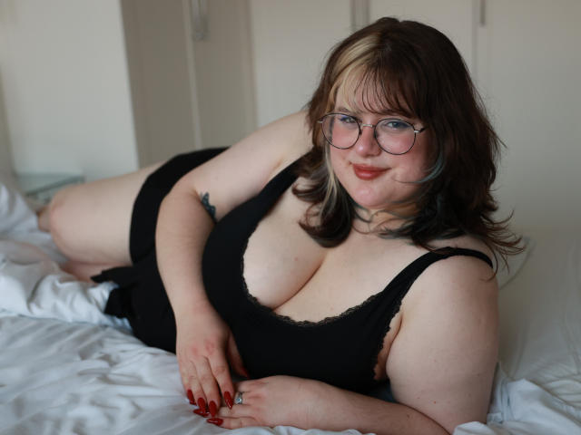 OpheliaLush, a plus-sized woman with glasses, red nails, and blonde highlights in brown hair, is lying on a bed in black lingerie, showing her big boobs.