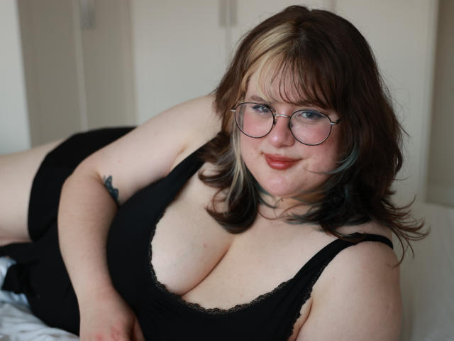 OpheliaLush with big tits in a black lace-trimmed tank top, lying on a bed, wearing glasses, has fair skin, brown hair with blonde highlights, and a tattoo on her arm.