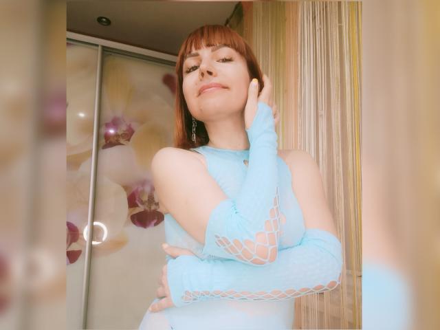 LaylaHottyX with light brown hair, blue fishnet dress, and matching sleeves, touching her cheek, smiling, showing small tits, in a softly lit room.