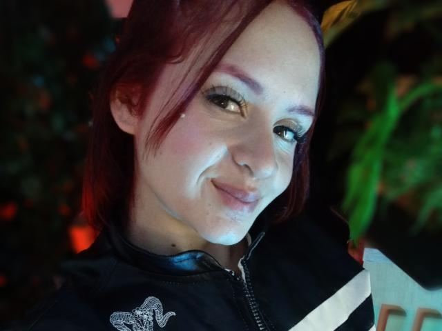 LesleyGray with red hair, light skin, and a small nose piercing, smiling slightly, wearing a black zip-up with a silver emblem, holding a white strap.