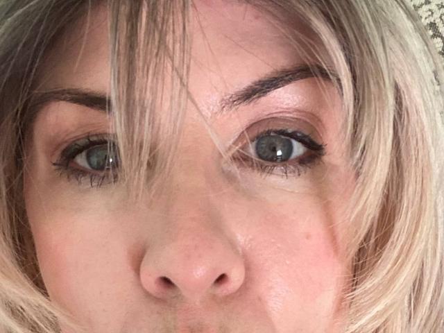 TianaQLove's face with blonde hair, blue eyes, and slightly parted lips, showing a small mole above her left nostril. Her eyebrows are thick and well-groomed.