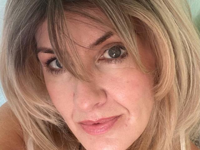 TianaQLove's face with blonde hair, green eyes, slightly wavy bangs, and a subtle smile, showing a small pimple on her right cheek. Her pink lips are slightly parted.