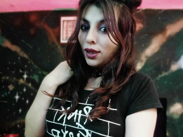 SoyVeronica hat dunkelbraunes Haar in lockigen Wellen, lila Lidschatten, schwarzen Lippenstift und trägt ein schwarzes Hemd mit weißer Schrift, berührt ihr Haar und hat eine schwarze Halskette..