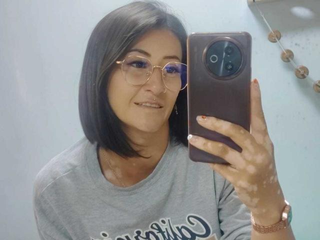 SharissWilliam, avec ses cheveux noirs mi-longs, ses lunettes et son anneau de nez, tenant un téléphone, portant une chemise grise, souriant, montrant des ongles sales et une montre rose..