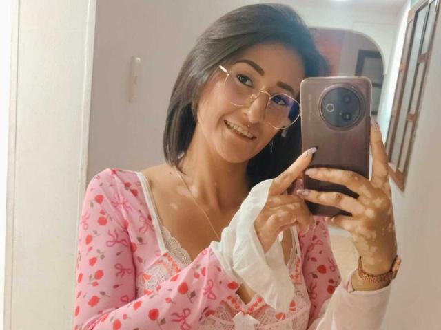 SharissWilliam, con gafas, sonriendo, sosteniendo un teléfono, vistiendo una camisa rosa con estampados rojos, mostrando sus pechos en un selfie frente a un espejo, sosteniendo un paño blanco con la otra mano..