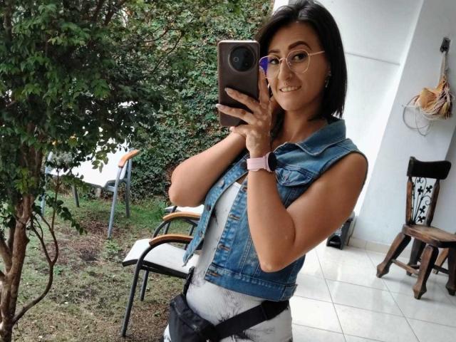 SharissWilliam with medium tits, wearing a denim sleeveless vest, white top, black belt, and pink watch, taking a selfie with glasses, outside on a tiled patio.