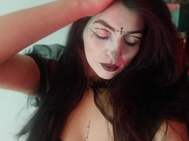 SoyVeronica met bleke make-up, donkere lipstick en zwarte eyeliner, hoofd naar beneden gekanteld, arm omhoog, lang zwart haar, kleine tatoeage onder de blote borsten, draagt een zwarte top..