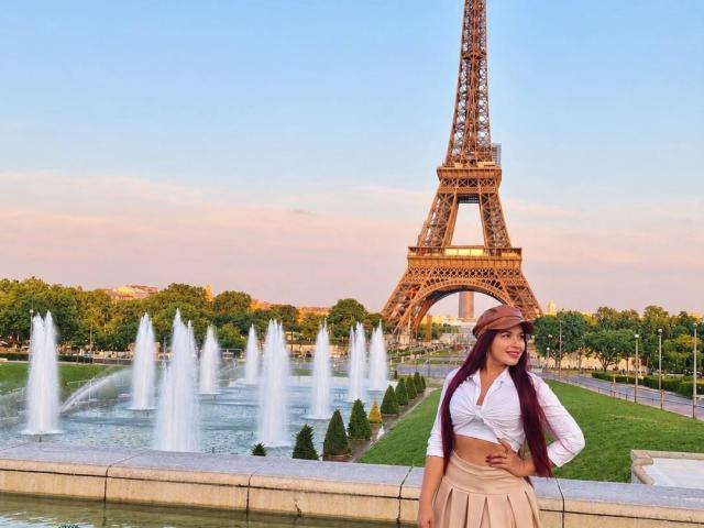 BigAssJessi con lunghi capelli viola, una camicia bianca annodata, una gonna beige e un cappello marrone, in piedi di fronte alla Torre Eiffel, con fontane d'acqua sullo sfondo..