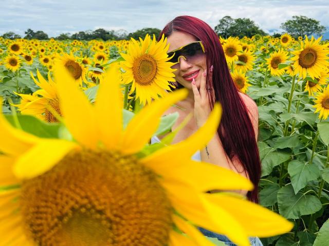 BigAssJessi con lunghi capelli rossi, occhiali da sole gialli, che sorride, toccando i petali di un girasole, in piedi in un campo di girasoli, mostrando il seno, girasoli a fuoco, cielo nuvoloso sullo sfondo..