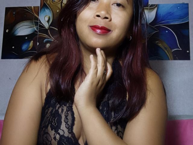 SarahFontaineX with red lipstick, black lace top, dark red hair, touching her chin, smiling, brown skin, medium tits, hoop earrings, blue and white floral wall art.