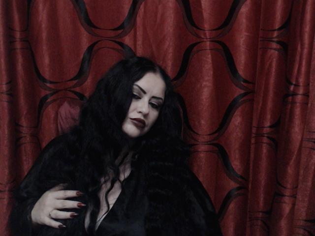 MssBlackWidow with long black hair, dark red lipstick, and black fur, holding her tits, against red wavy-patterned curtains, with red-painted nails. She's looking down, seductive.